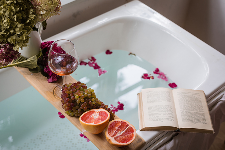 Bath tub with a tray with grapefruit slices, bunch of grapes, a glass of wine and a book | Innovate Building Solutions | #Bathtube #ShowerTub #SpaDay #BathroomSpa